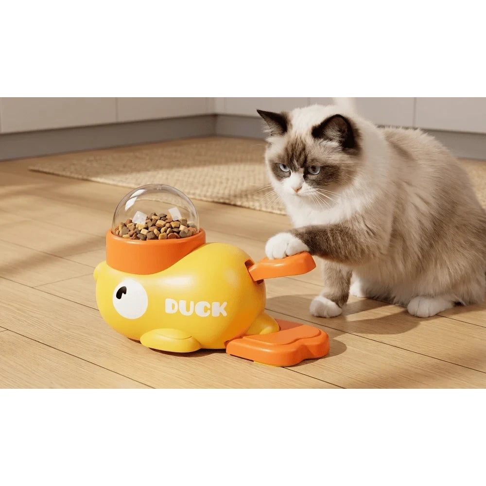 Cat beside a yellow duck-shaped manual treat dispenser on a wood surface.