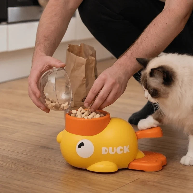Person adding treats to the open yellow duck-shaped treat dispenser while a cat watches.